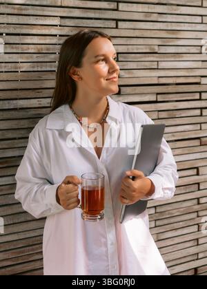 Eine junge Frau, die mit einem Laptop bleibt und eine Tasse Tee hält Stockfoto