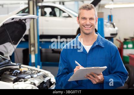 Männlicher Mechaniker lächelt, hält Klemmbrett und Stift in der Werkstatt neben dem offenen Motorraum Stockfoto