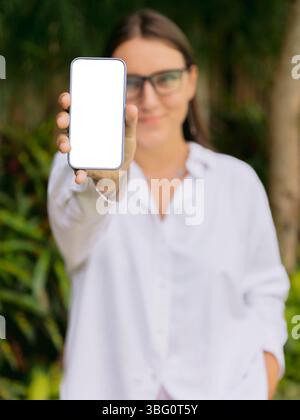 Lächelnde Frau zeigt ein Smartphone mit leerem Bildschirm draußen. Mockup für App-, Website- oder Social-Media-Design. Stockfoto