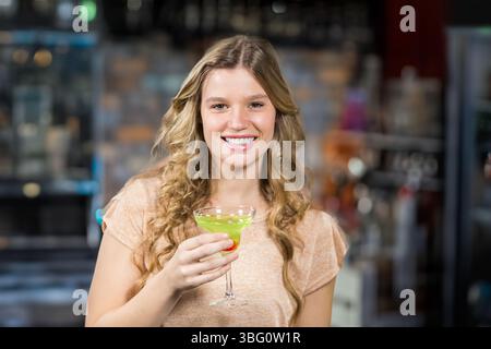 Frau in ihren Zwanzigern lächelt, während sie einen grünen Cocktail in Martini-Glas an der Bar hält Stockfoto