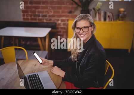 Frau mit Brille und Lederjacke, die am Tisch im Café sitzt und Smartphone und Laptop hält Stockfoto