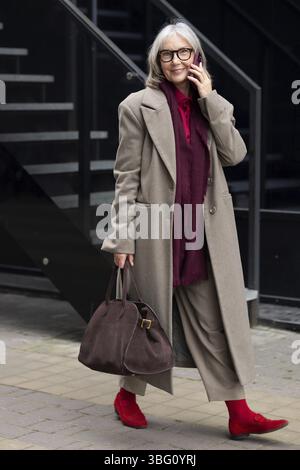 Eine ältere Geschäftsfrau in einem schicken grauen Mantel und knallroten Schuhen läuft selbstbewusst, während sie am Telefon in der Nähe zeitgenössischer Gebäude spricht Stockfoto