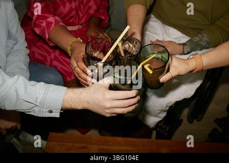 Vielfältige Gruppe von jungen Erwachsenen und Menschen mittleren Alters mit Behinderungen klirrrend Brille während der Geburtstagsfeier, sichtbare Hände mit unterschiedlichen Hauttönen und Rollstuhlaufnahmen Stockfoto
