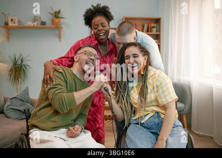 Verschiedene Gruppen junger Erwachsener feiern Geburtstag, kaukasischer Mann, der im Rollstuhl ins Mikrofon singt, während Freunde gemeinsam im Wohnzimmer Spaß haben Stockfoto