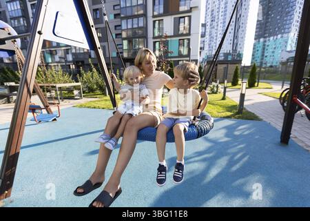 Eine Mutter engagiert sich freudig mit ihren beiden Söhnen, während sie zusammen auf einer Schaukel auf einem lebhaften Spielplatz spielen. Die Gegend ist von modernen Gebäuden umgeben, Indi Stockfoto