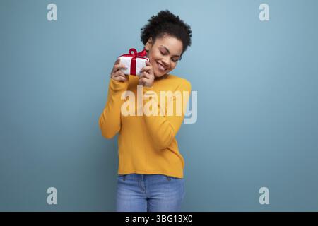Fröhliche junge brünette lateinamerikanische Erwachsene in gelbem Pullover erhielt ein Geschenk in einer Box Stockfoto