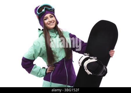 Isoliert auf weißem, attraktivem weißem Mädchen in lila Ski-Outfit, roter Hose und lila Wollmütze, schwarzem Snowboard halten, in die Kamera schauen, lächeln, ha Stockfoto