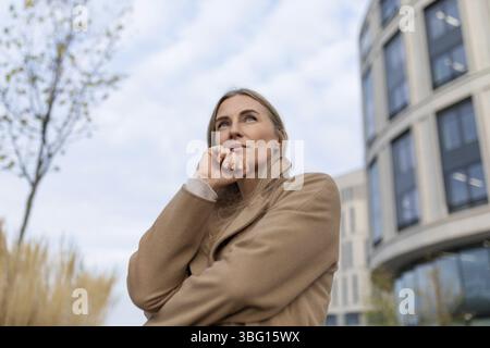 Eine Frau mit blonden Haaren steht vor einem Business Center. Sie trägt einen langen, beigefarbenen Mantel und blickt nachdenklich auf Stockfoto