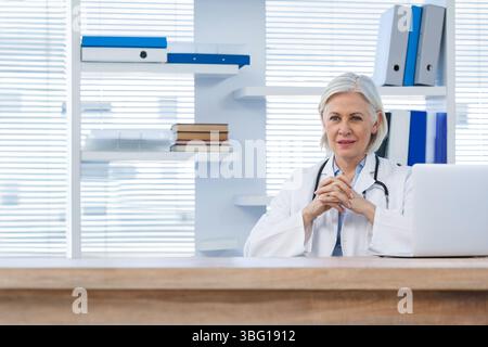 Leitende Ärztin sitzt am Schreibtisch in der Klinik mit Laptop und Stethoskop, Kopierraum Stockfoto