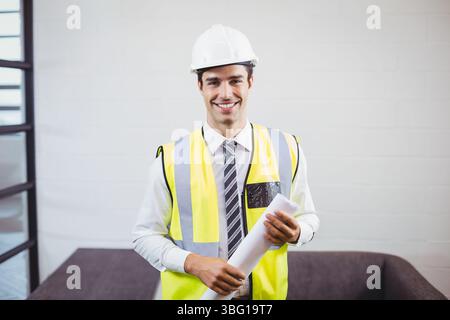 Bauingenieur trägt weißen Schutzhelm und gelbe Sicherheitsweste und hält Pläne im Büro Stockfoto