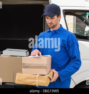 Männlicher Kurier in Uniform, der die Zwischenablage überprüft und Pakete neben dem weißen Van auf einer gepflasterten Einfahrt hält Stockfoto