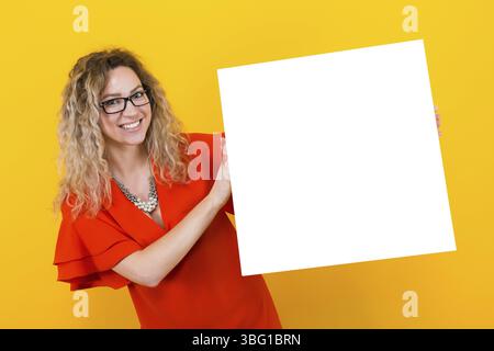 Porträt der attraktiven glücklichen lockigen Frau in rotem Kleid und Brille isoliert auf orangem Hintergrund, die emty leeres Brett hält Ihr Text hier conc Stockfoto