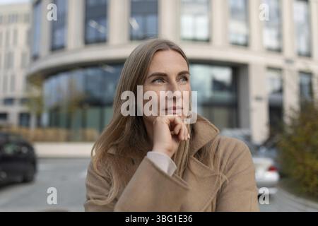 Eine junge Frau mit langen blonden Haaren steht vor einem Business Center. Sie trägt einen beigefarbenen Mantel und blickt nachdenklich nach rechts o Stockfoto