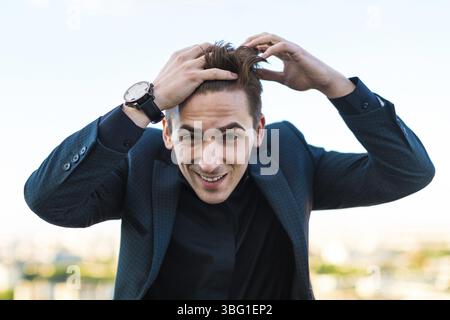 Junger gut aussehender brünette kaukasischer Geschäftsmann in dunklem Anzug, schwarzem Hemd und teurem Uhrenständer auf dem Dach, Blick in die Kamera, lächelnd, begradigt h Stockfoto