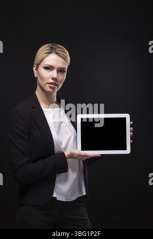 Erfolgreiche junge Geschäftsfrau in einem stilvollen schwarzen Business-Anzug und einer weißen Bluse auf schwarzem Hintergrund, die das Tablet-Display selbstbewusst nach vorne hält Stockfoto