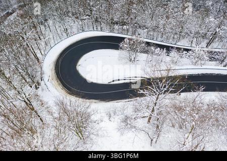Aus der Vogelperspektive auf eine kurvenreiche schwarze Asphaltstraße, die sich durch eine schneebedeckte Waldlandschaft schlängelt Stockfoto