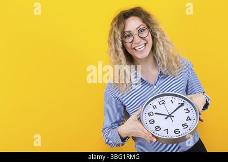 Porträt einer lächelnden Frau mit lockigen Haaren in gestreiftem Hemd und runder Brille isoliert auf orangem Hintergrund, die Uhren hält Zeitlimit Pünktlichkeit Conce Stockfoto