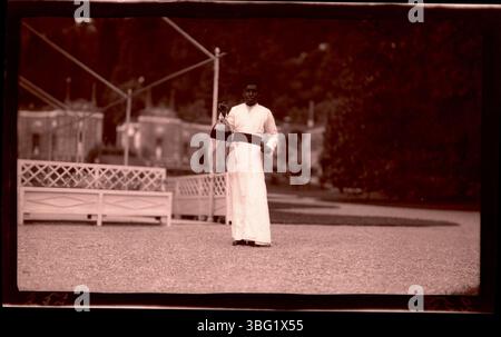 1913 wird ein Mann in einem weißen Gewand und fès gesehen, der vor dem Mosaikhaus in der Villa d’Este am Comer See einen Dinner gong hält. Die Arrases besuchten die Villa während ihrer Europa-Tour und nahmen dieses Foto auf, um ihre Erfahrungen an diesem berühmten italienischen Wahrzeichen zu dokumentieren. Stockfoto