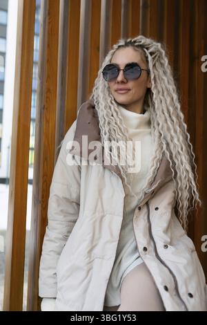 Eine Frau mit langen blonden Haaren wird mit einem weißen Mantel gesehen. Sie steht selbstbewusst und zeigt ihre elegante Kleidung mit Pose und Anmut Stockfoto