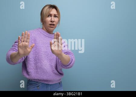 Porträt einer entrüsteten blonden jungen Frau in einem Lavendelpullover mit einem Zeichen der Verleugnung auf einem hellen, glatten Hintergrund Stockfoto