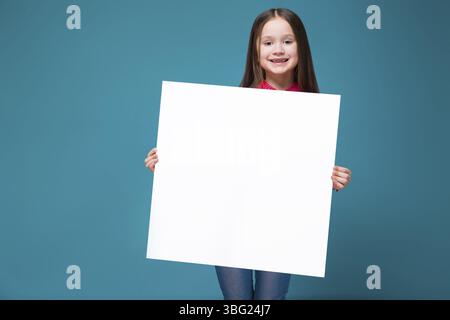 Isoliert auf blauem, wunderschönem weißbrünetten Kind in rotem kariertem Hemd und blauer Jeans mit langen Haaren halten leeres Poster, schauen Sie in die Kamera, stehen Sie dahinter Stockfoto