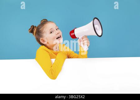 Isoliert auf blauem, entzückendem rothaarigem Mädchen mit Sommersprossen in Zitronenpullover mit weißem Megaphon, der in die Kamera blickt. Die Hände sind über dem Weißen Stockfoto