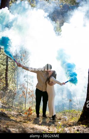 Fröhlich und fröhlich feiert ihr Schwangerschaftspaar, umgeben von blauem Rauch in einem wunderschönen, üppigen Wald Stockfoto