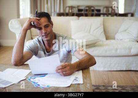 Mann, der auf dem Boden im Wohnzimmer sitzt, mit einem Stift, der Kartenpapiere an einem niedrigen Couchtisch überprüft Stockfoto