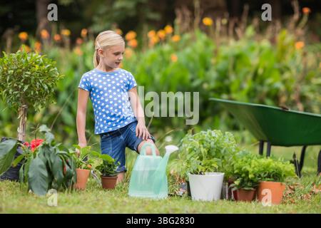 Mädchen, das mit dem Fuß auf Topfpflanzen in der Nähe von Gießkanne und Schubkarre im Garten steht Stockfoto
