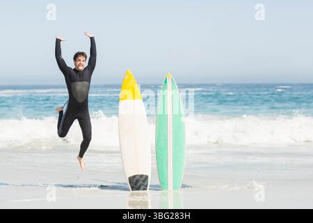 Mann in schwarzem Neoprenanzug, der an der Küste neben weißen und grünen Surfbrettern springt, Kopierraum Stockfoto