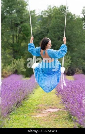 Iwano-Frankiwsk, Ukraine 18. Juli 2024: Eine junge Frau in einem hellblauen Kleid sitzt auf einer weißen Schaukel in einem malerischen Lavendelfeld. Die Szene evo Stockfoto