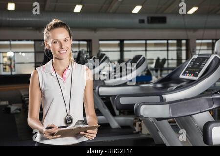 Weibliche Fitnesstrainerin lächelt neben Laufbändern im Fitnessstudio und hält Klemmbrett und Stift mit Stoppuhr Stockfoto