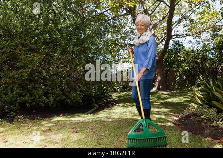 Gartenrechen, der gefallene Blätter auf Grasrasen in der Nähe von geschwungenem Blumenbeet unter strahlendem Sonnenschein schwärzt Stockfoto