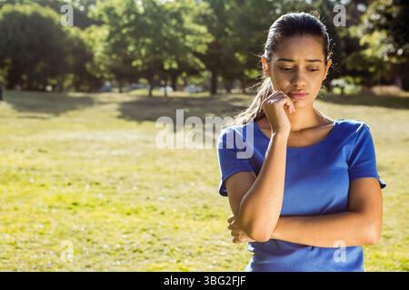 Asiatische Frau, die im sonnendurchfluteten Park steht, das Kinn auf der Hand ruht, während sie Grasfelder, Kopierraum vermisst Stockfoto