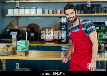 Männlicher Barista lehnt sich auf Holztheke im Café mit Brotkorb, Espresso-Maschine und Kopierraum Stockfoto