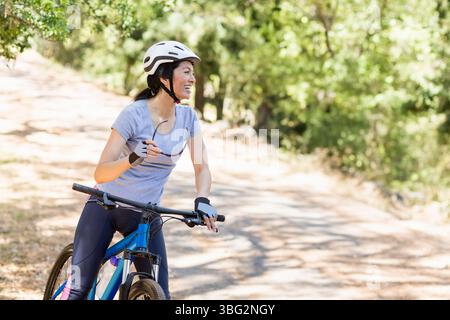 Asiatische Frau, die auf dem sonnendurchfluteten Waldweg eine Sonnenbrille hält, Helm und Fahrrad trägt Stockfoto