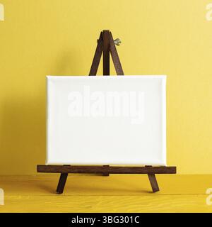 Staffelei mit leerer Leinwand auf Holztisch. Gelber Wandhintergrund Stockfoto