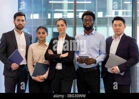 Eine vielfältige Gruppe von Geschäftsleuten, die zusammen in einem modernen Büroumfeld stehen. Sie blicken direkt auf den Zuschauer, mit einem selbstbewussten Verhalten. Stockfoto