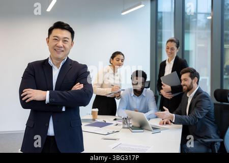 Ein selbstbewusster asiatischer Geschäftsmann lächelt mit überkreuzten Armen und leitet ein vielfältiges Team in einer modernen Büroumgebung. Stockfoto