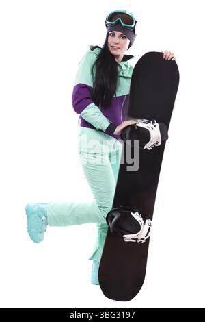 Vertikales Bild, isoliert auf weißer, hübscher kaukasischer Frau in lila Ski-Outfit und blauen Hosen halten schwarzes Snowboard, Bein hoch, Blick in die Kamera Stockfoto