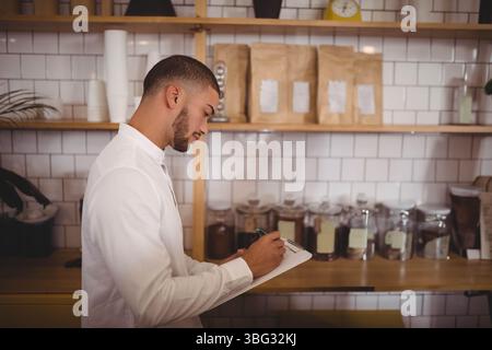 Männlicher Barista schreibt auf die Zwischenablage an der Theke mit beschrifteten Glasgläsern und Pappbechern, Kopierraum Stockfoto