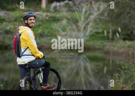 Mann lächelt auf dem Mountainbike am Rand des Waldteichs mit Regenjacke und Helm, Kopierraum Stockfoto
