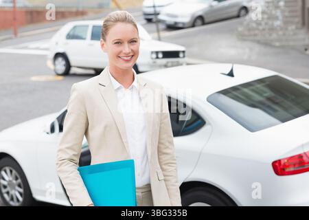 Geschäftsfrau, die neben weißer Limousine und Fließheck auf der städtischen Straße steht, hält blaue Mappe lächelnd Stockfoto