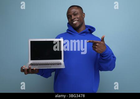Hübscher afrikanischer junger Berater in stilvollem Hoodie mit Laptop, um Werbeanzeigen im Hintergrund mit Kopierraum zu zeigen Stockfoto