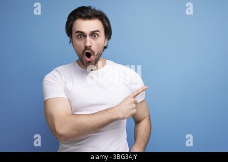 Schockiert von einem überraschenden, brünetten jungen Mann in einem weißen T-Shirt mit Bizeps auf einem Studio-Hintergrund mit Copyspace Stockfoto
