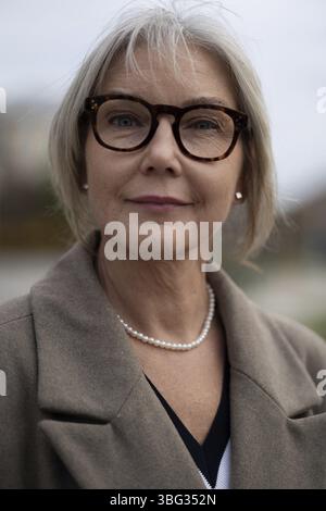 Eine reife Geschäftsfrau steht draußen und sieht geschickt und selbstbewusst aus. Sie trägt einen schicken Mantel und eine stilvolle Brille, wobei die sanfte natürliche Beleuchtung h Stockfoto
