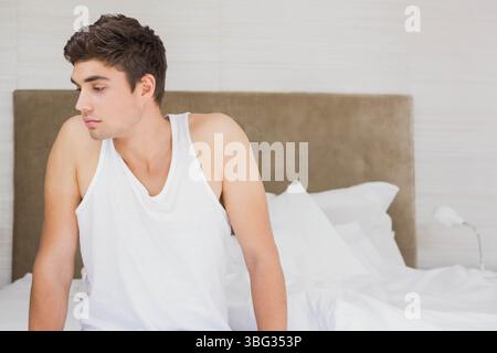 Mann in seinen Zwanzigern, der auf der Bettkante sitzt, in sanft beleuchtetem Schlafzimmer mit kleiner Nachttischlampe Stockfoto
