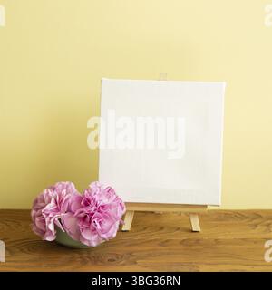 Staffelei und leere Leinwand mit Nelkenblumen auf Holztisch. Gelber Hintergrund Stockfoto