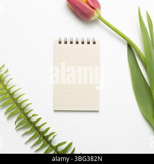 Leerer Notizblock mit Tulpenblume auf weißem Hintergrund. Blumendesign, flacher Lay, Draufsicht, Kopierbereich Stockfoto