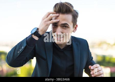 Junger, gut aussehender brünette Geschäftsmann in dunklem Anzug, schwarzem Hemd und teurem Uhrenständer auf dem Dach, Blick in die Kamera, lächelnd, Hand auf Gesicht Stockfoto